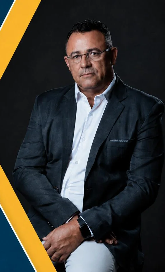 Douglas Diniz, de paletó escuro e camisa branca, sentado, em fundo escuro